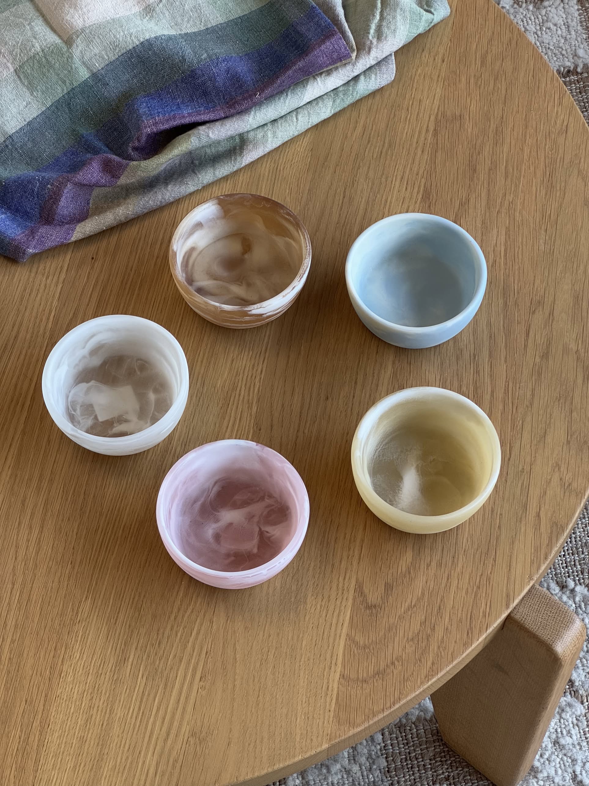 Five small, round HKLIVING Patio Bowl cups in pastel marbled resin (brown, blue, white, pink, and yellow) are arranged on a wooden table with a folded plaid cloth partly visible—perfect for outdoor entertaining.