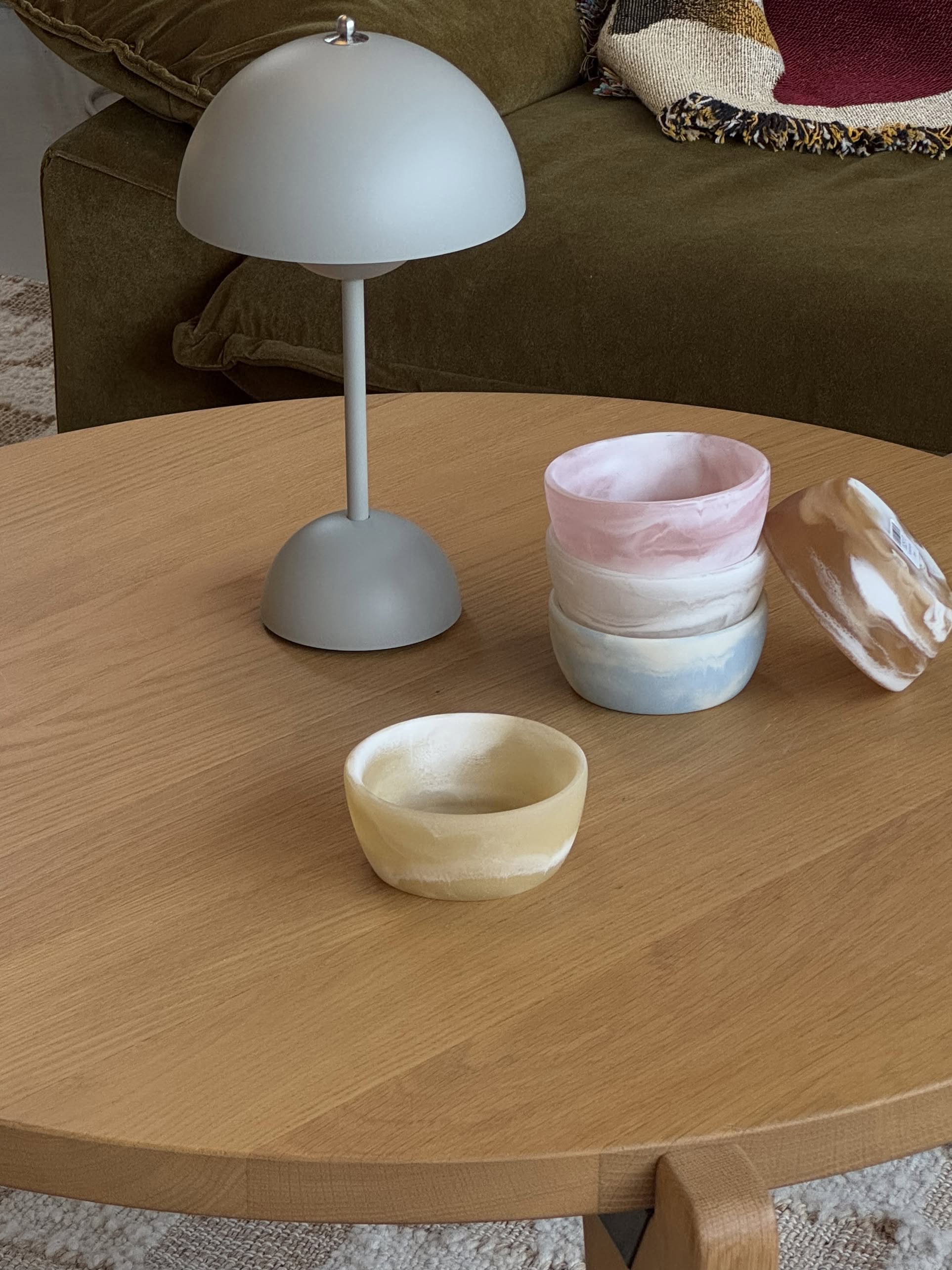 A round wooden coffee table with a gray dome lamp displays five HKLIVING Patio Bowls by HKLIVING in pastel pink, blue, yellow, and beige, beautifully stacked in front of an olive green couch—ideal for outdoor entertaining.