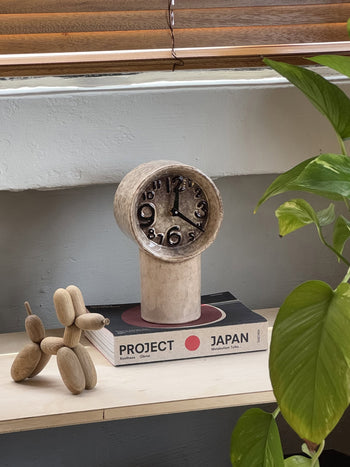 The HKLIVING Retro Ceramic Clock - Cream sits on a Japan-themed book beside a small wooden balloon dog and a leafy green plant, all beneath a window with wooden blinds.