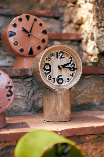 The HKLIVING Retro Ceramic Clock - Cream stands on a stone step outdoors, surrounded by other quirky, handmade HKLIVING clocks. Sunlight casts playful shadows over the vintage-inspired scene.