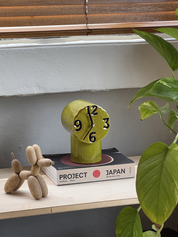 The HKLIVING Retro Ceramic Clock – Olive, a wooden balloon dog sculpture, and the book Project Japan sit on a light wooden shelf beside a leafy plant by the window, creating a vignette of vintage-inspired design.