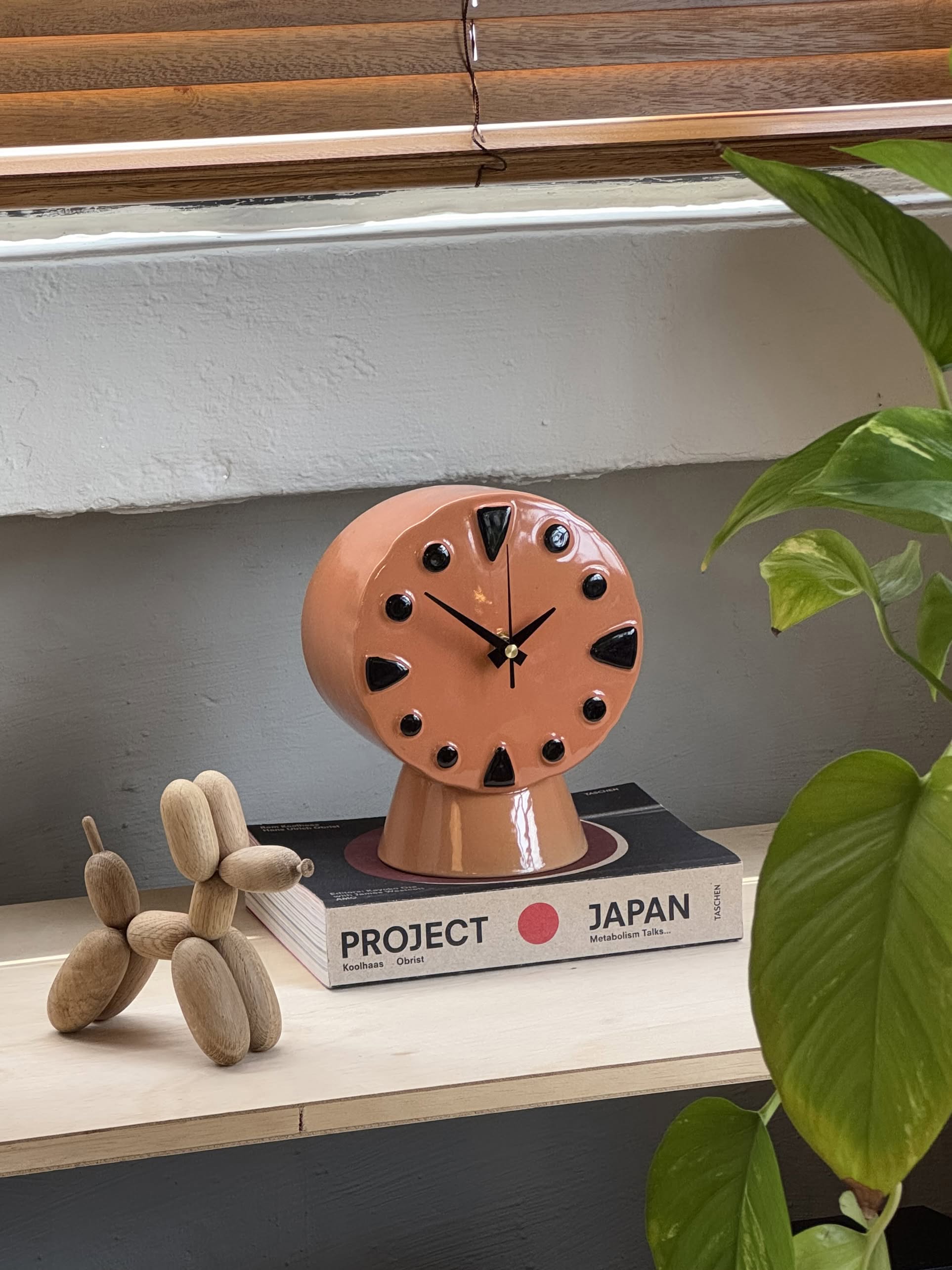 The HKLIVING Retro Ceramic Clock - Peach, with black dots and triangle markers, rests on a Project Japan book atop a shelf beside a wooden dog sculpture and green plant, all illuminated by sunlight.