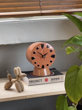 The HKLIVING Retro Ceramic Clock - Peach, with black dots and triangle markers, rests on a Project Japan book atop a shelf beside a wooden dog sculpture and green plant, all illuminated by sunlight.