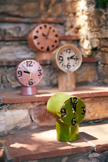 The HKLIVING Retro Ceramic Clock – Peach, by HKLIVING, features a vintage-inspired design with distorted shapes and numbers in soft hues. Displayed on outdoor stone steps, its ceramic body glows in the sunlight for a stylish retro look.