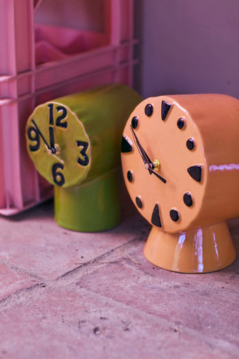 Two HKLIVING Retro Ceramic Clocks—one green with bold black numbers, one peach with triangle markers—crafted using traditional firing methods, rest on a tiled floor beside a pink crate.