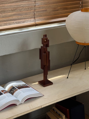 The HKLIVING Skyline Sculpture S - Espresso sits on a light wood shelf beside an open book and a round white lamp with black tripod legs. Above, horizontal blinds call to mind urban architecture.