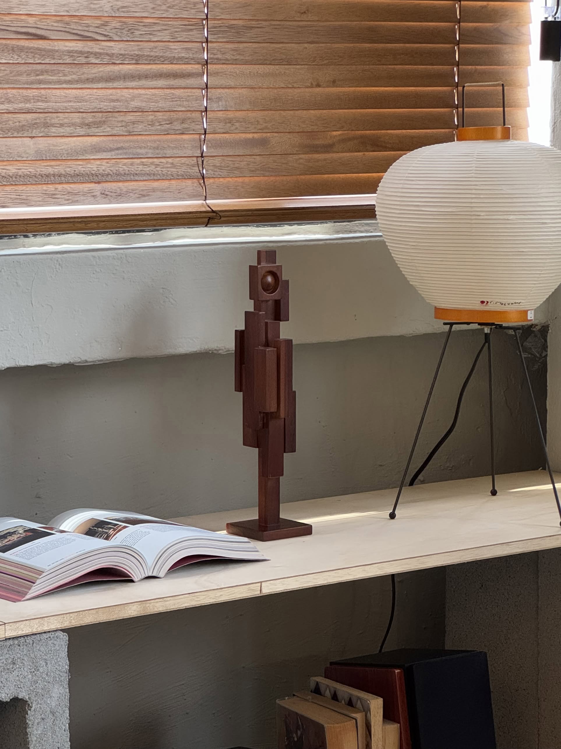 A teak wood shelf holds an open book, the HKLIVING Skyline Sculpture S - Espresso, and a paper lantern lamp. Wooden blinds cover the window above, with book spines visible on a lower shelf.