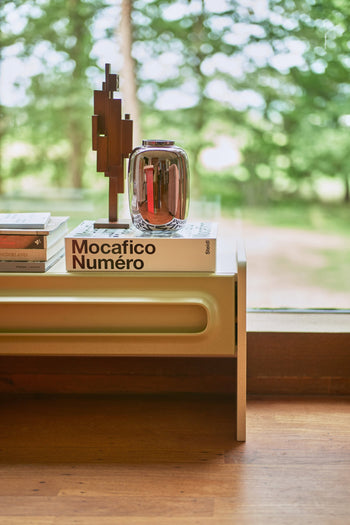 A modern teak wood coffee table by a large window displays stacked books, including one titled Mocafico Numéro, a shiny glass vase, and the HKLIVING Skyline Sculpture S - Espresso. Lush greenery can be seen outside.