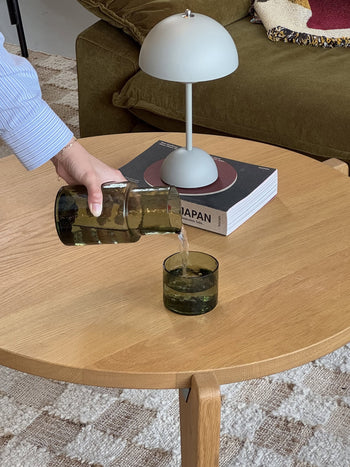 A hand pours water from the HKLIVING Tube Carafe & Glass Set - Olive into a matching glass on a round wooden table, with a lamp and the book JAPAN in the background—ideal for contemporary dining.