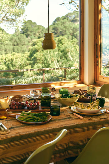 A sunlit dining table for contemporary dining features plates of asparagus, grapes, bread, and salad alongside the HKLIVING Tube Carafe & Glass Set - Olive by HKLIVING. A large window overlooks lush green trees and hills for a warm, inviting atmosphere.