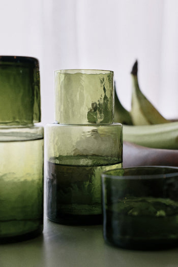 The HKLIVING Tube Carafe & Glass Set - Olive features stylish green glassware with a textured finish, perfect for modern dining, displayed on a surface with blurred bananas and a softly lit background.