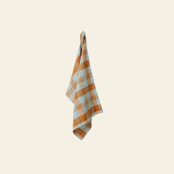 The HKLIVING Vida Check Tea Towel in Amber/Blue features orange and light blue checks, hanging on a hook against a light beige background—perfect for adding color to your kitchen decor.