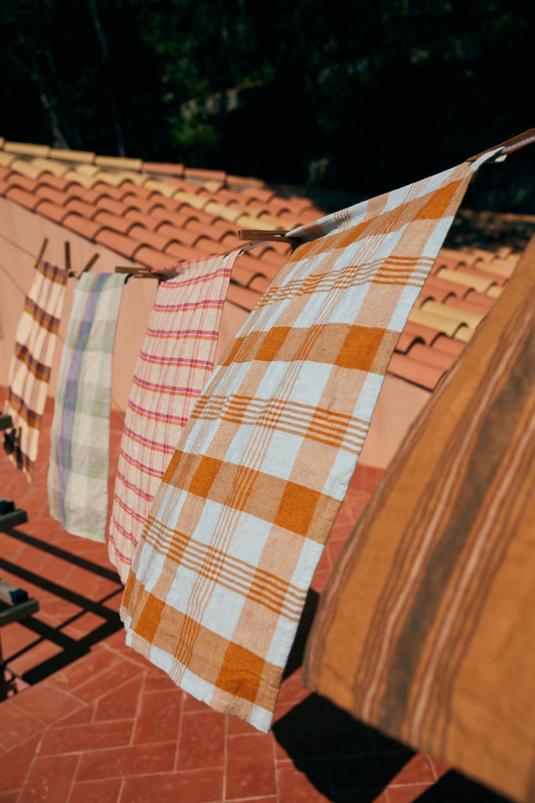 The HKLIVING Vida Check Tea Towel in Amber/Blue hangs on a clothesline above a terracotta-tiled patio, with a red-tiled roof and trees in the background.