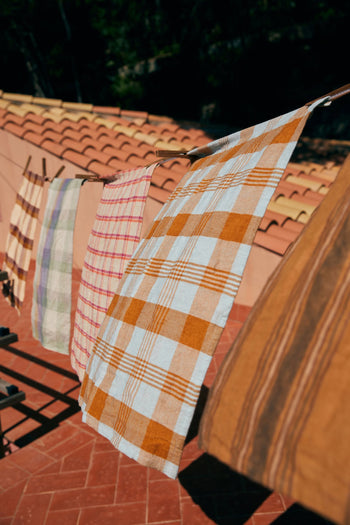 The HKLIVING Vida Check Tea Towel in Amber/Blue hangs on a clothesline above a terracotta-tiled patio, with a red-tiled roof and trees in the background.