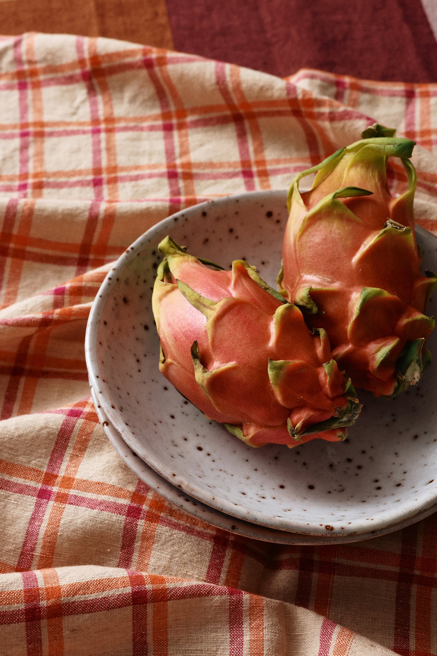 Two ripe dragon fruits with pink skin and green tips rest on a white speckled ceramic plate, placed atop the HKLIVING Vida Check Tea Towel in Strawberry/Cream by HKLIVING.