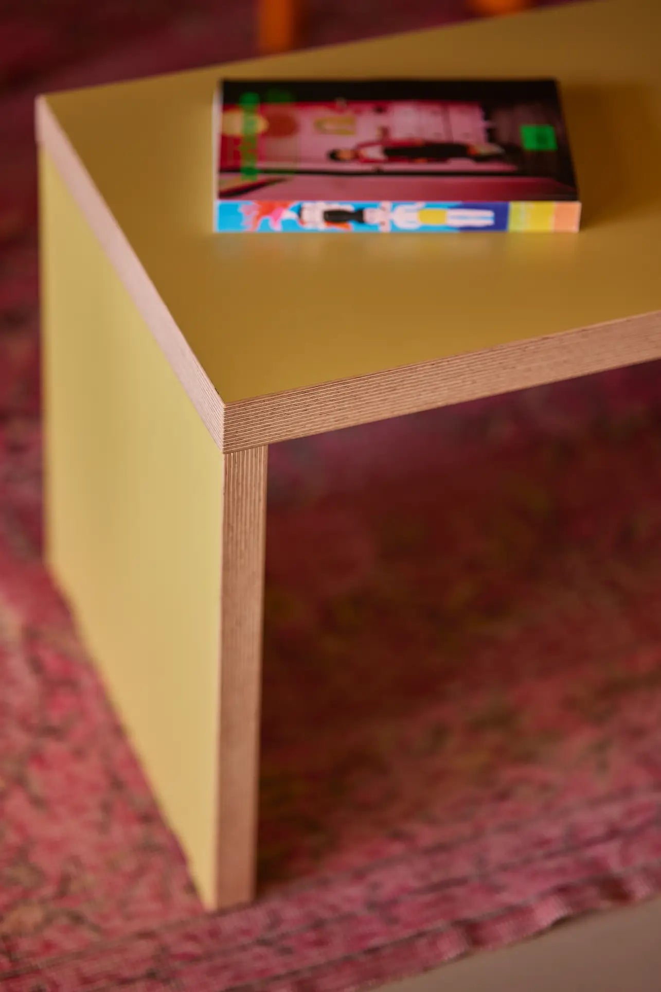 A close-up of a modern yellow wooden table with a colorful book atop, set on a pink patterned rug next to the versatile HKLIVING Bench, 180cm by HKLIVING, offering extra seating.