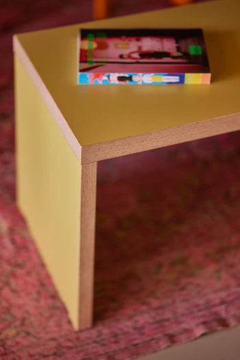 A close-up of a modern yellow wooden table with a colorful book atop, set on a pink patterned rug next to the versatile HKLIVING Bench, 180cm by HKLIVING, offering extra seating.