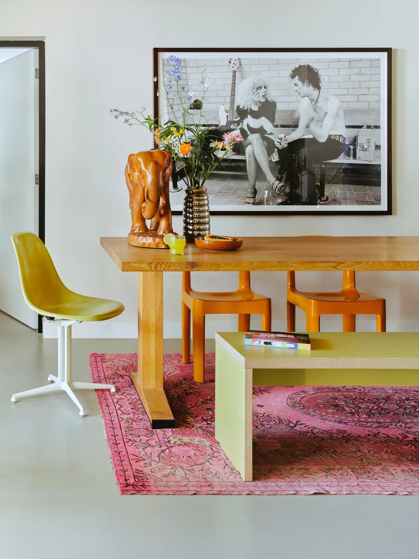 A modern dining area with a wooden table, orange stools, a green HKLIVING Bench (180cm), a yellow swivel chair, and a pink rug. On the table are flowers in a vase and an elephant sculpture; a black-and-white photo hangs on the wall.