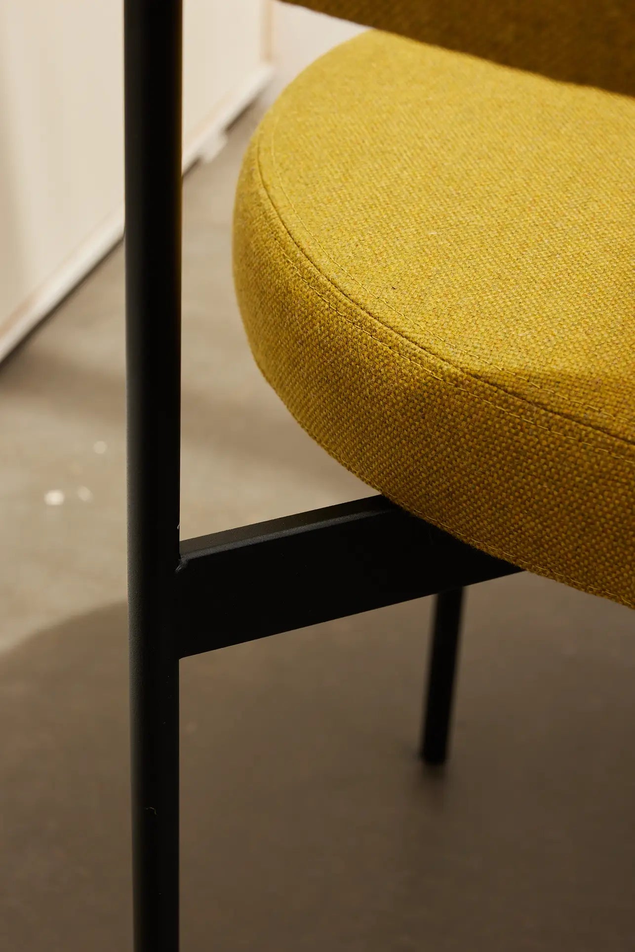 Close-up of the HKLIVING Dining Armchair in Main Line Flax with a round mustard yellow fabric seat and black metal legs, highlighting sleek Dutch design on a smooth gray floor.