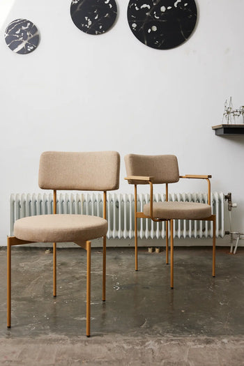 Two HKLIVING Dining Armchairs in Main Line Flax, Dusty by HKLIVING with gold metal frames sit before a white wall and radiator. Three round, abstract black and white artworks hang above, enhancing the modern look.