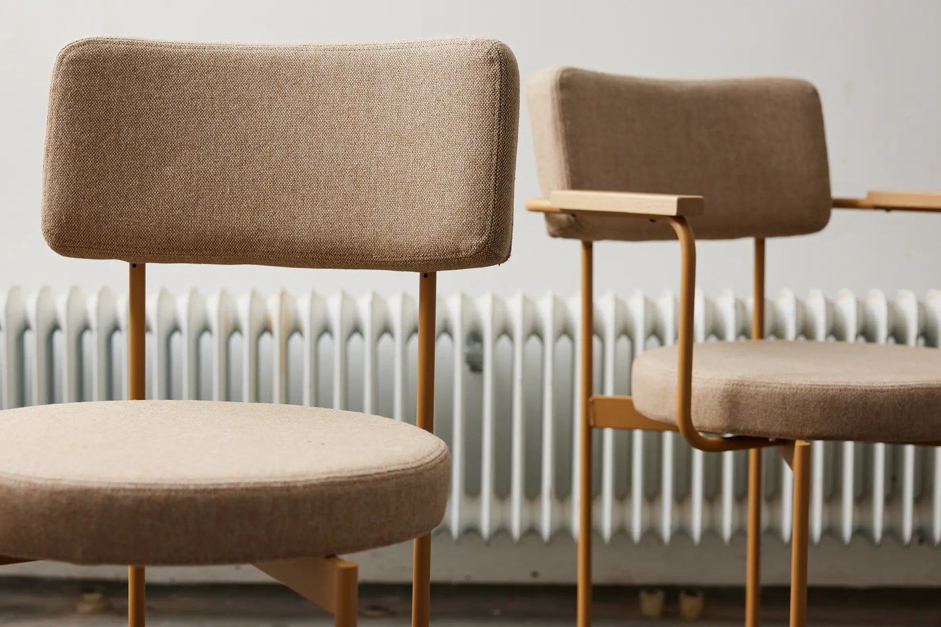 Two HKLIVING Dining Armchairs in Main Line Flax Dusty, featuring cushioned seats, metal legs, and armrests, add a minimalistic touch in front of a white radiator against a light-colored wall.
