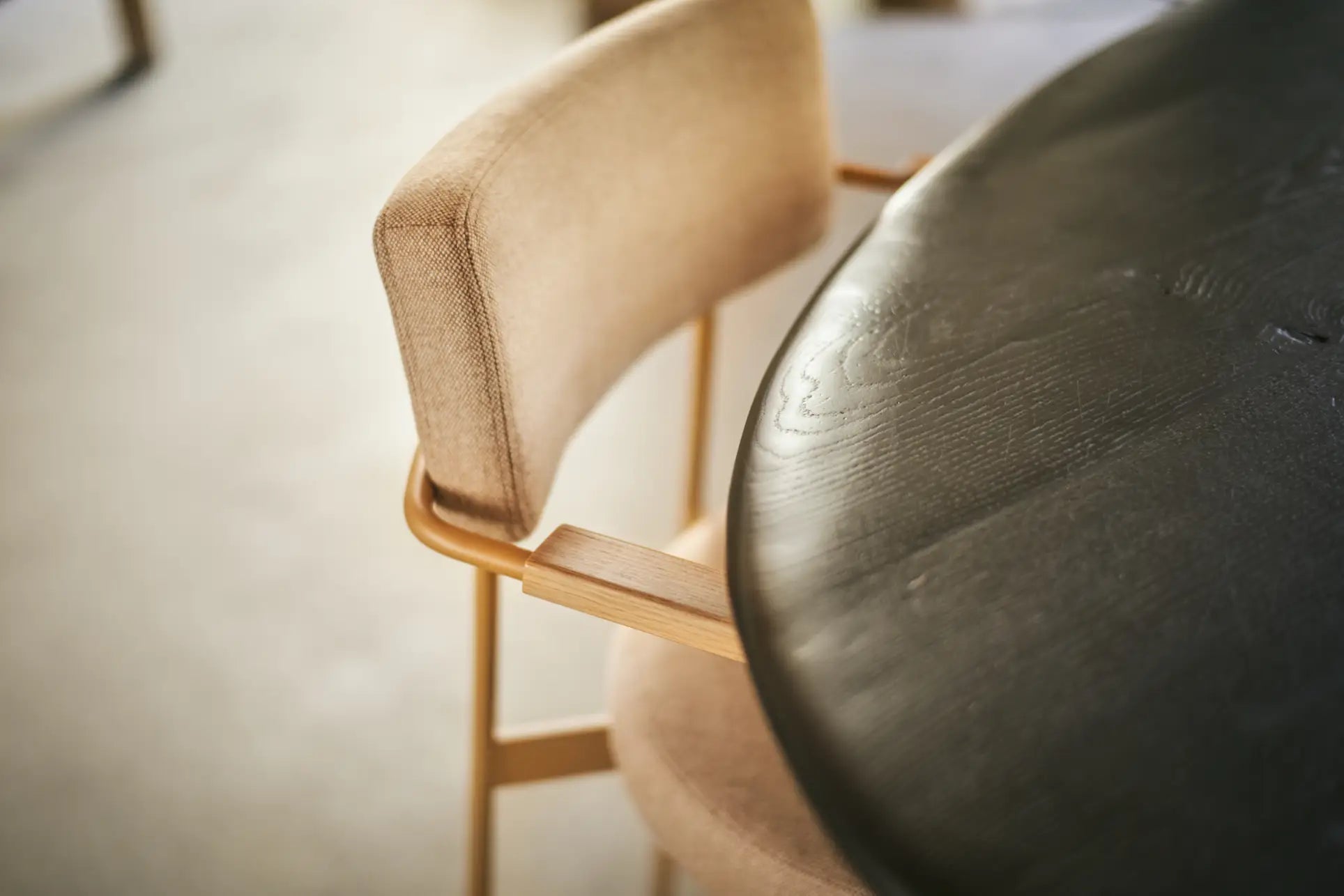 A close-up of the HKLIVING Dining Armchair, Main Line Flax Dusty, with light wooden legs beside a round dark wood table on a light carpeted floor.