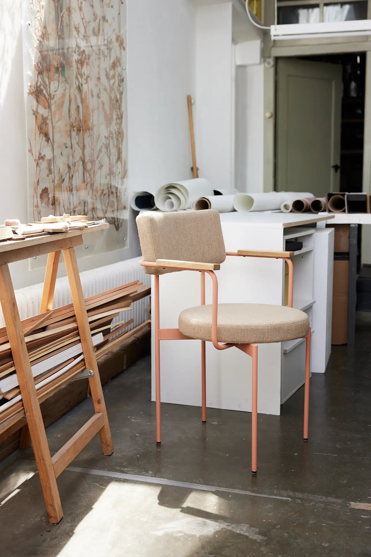 The HKLIVING Dining Armchair, Main Line Flax, Nude by HKLIVING features pink metal legs and is set in a bright studio with rolled-up papers, wooden planks, and a worktable by a window with floral curtains on a concrete floor.