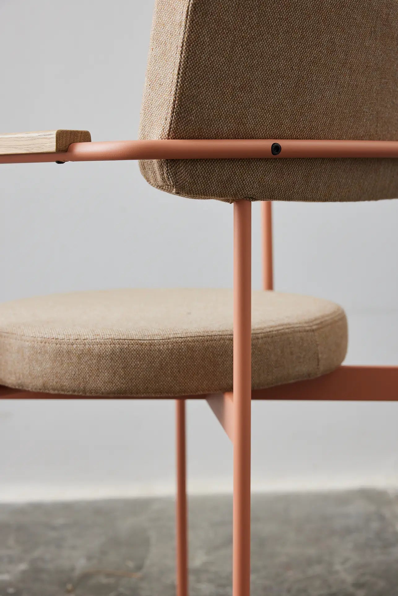 Close-up of the HKLIVING Dining Armchair, Main Line Flax, Nude by HKLIVING, featuring a beige upholstered seat and backrest with slim light pink metal legs and frame, shown against a plain gray wall and concrete floor.