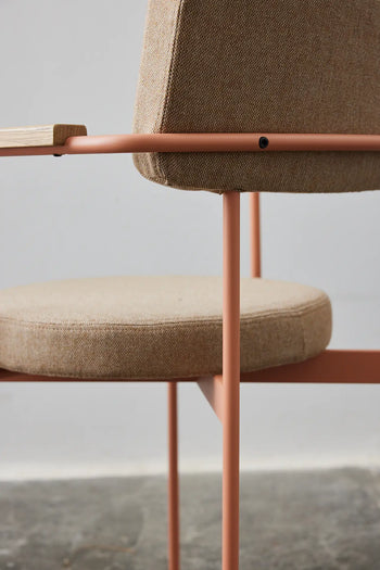 Close-up of the HKLIVING Dining Armchair, Main Line Flax, Nude by HKLIVING, featuring a beige upholstered seat and backrest with slim light pink metal legs and frame, shown against a plain gray wall and concrete floor.