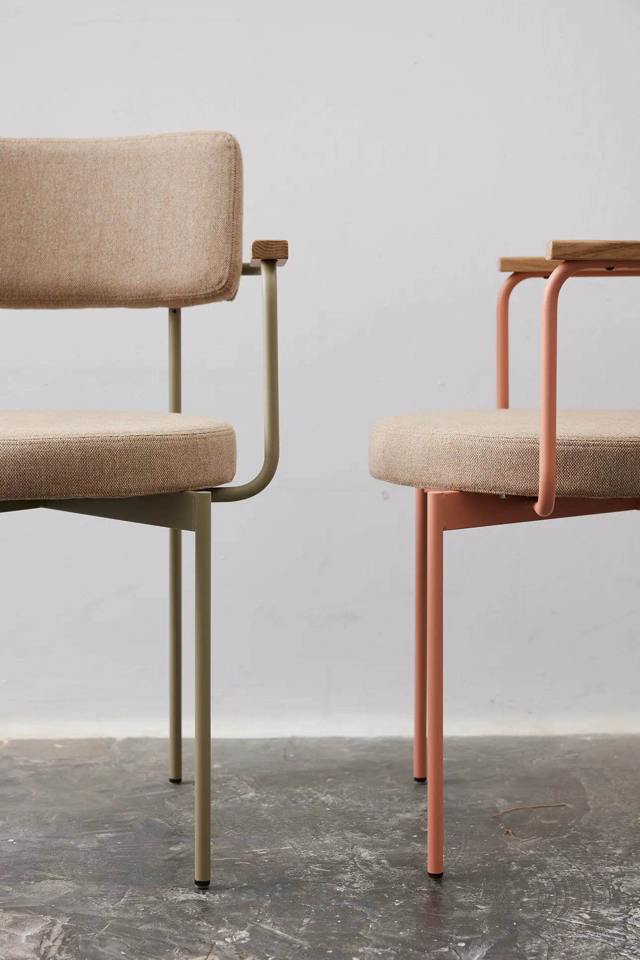 Two HKLIVING Dining Armchairs, Main Line Flax in Nude, featuring upholstered beige seats and backs—one with light green metal legs, the other with pink—are set on a gray concrete floor against a plain light wall.