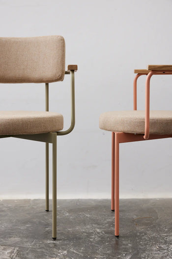 Two HKLIVING Dining Armchairs, Main Line Flax in Nude, featuring upholstered beige seats and backs—one with light green metal legs, the other with pink—are set on a gray concrete floor against a plain light wall.