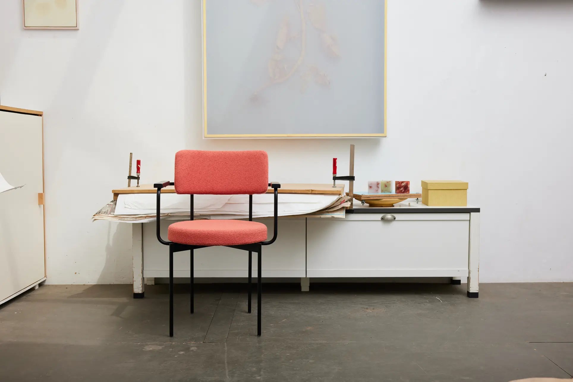An HKLIVING Dining Armchair, Yoredale, Black with plush cushioning and black metal legs is centered in front of a white desk in a minimalist room with Dutch design, white walls, and displayed art.