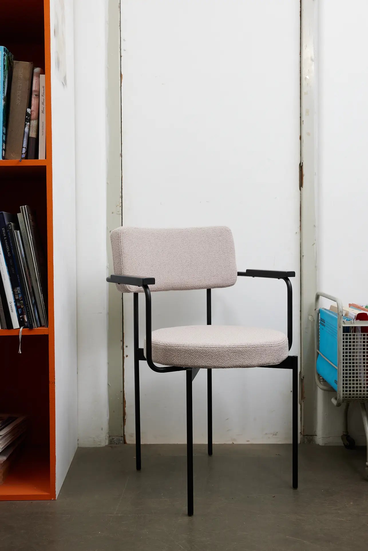 The HKLIVING Dining Armchair, Yoredale, Black features a customizable design with light beige cushions and a black metal frame. It sits on a gray floor between a bookshelf and a blue rolling cart.