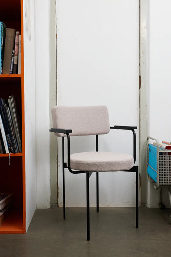 The HKLIVING Dining Armchair, Yoredale, Black features a customizable design with light beige cushions and a black metal frame. It sits on a gray floor between a bookshelf and a blue rolling cart.