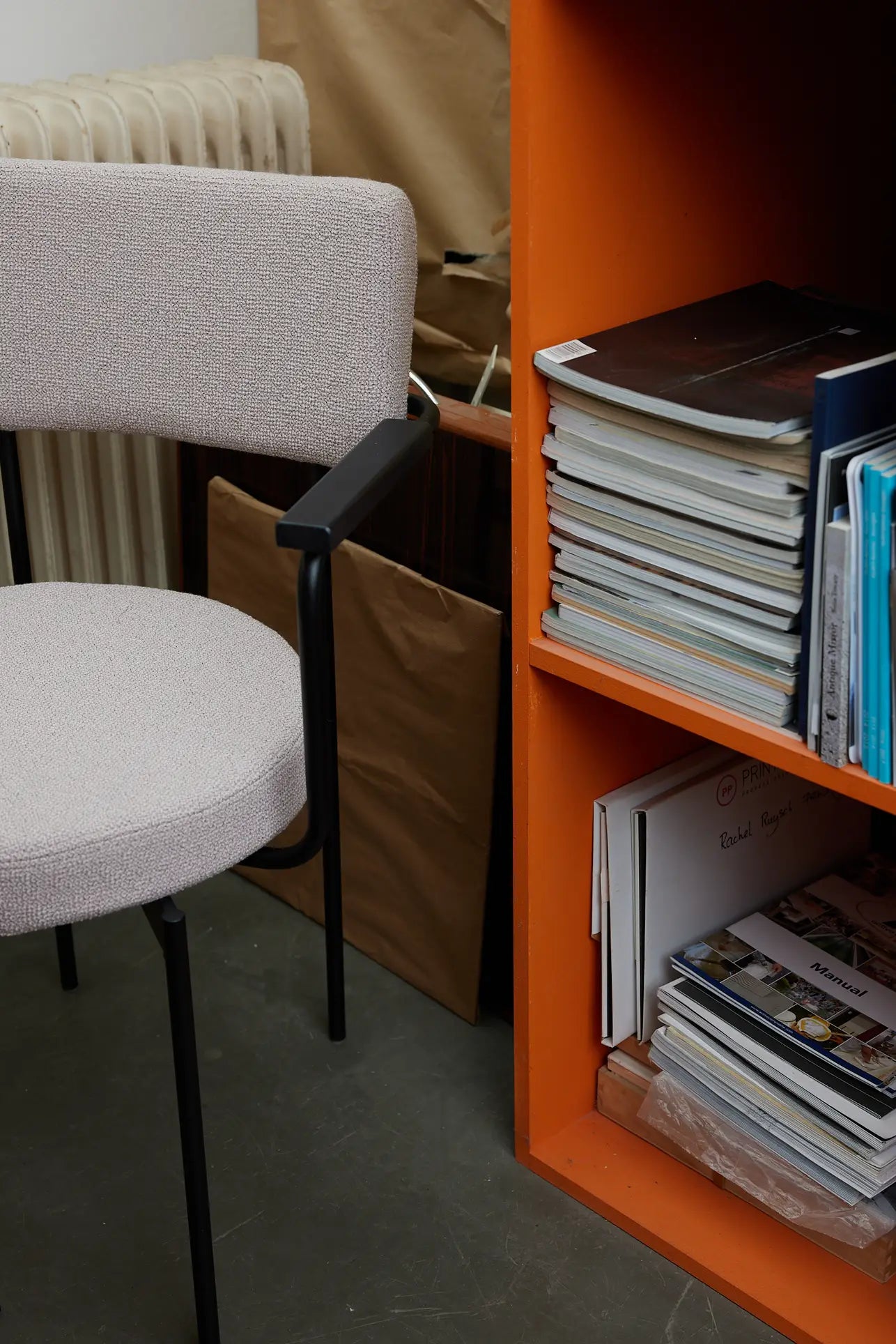 A black HKLIVING Dining Armchair, Yoredale, sits next to an orange bookshelf filled with magazines, books, and folders; a brown paper bag and a radiator are in the background of this Dutch-inspired scene.