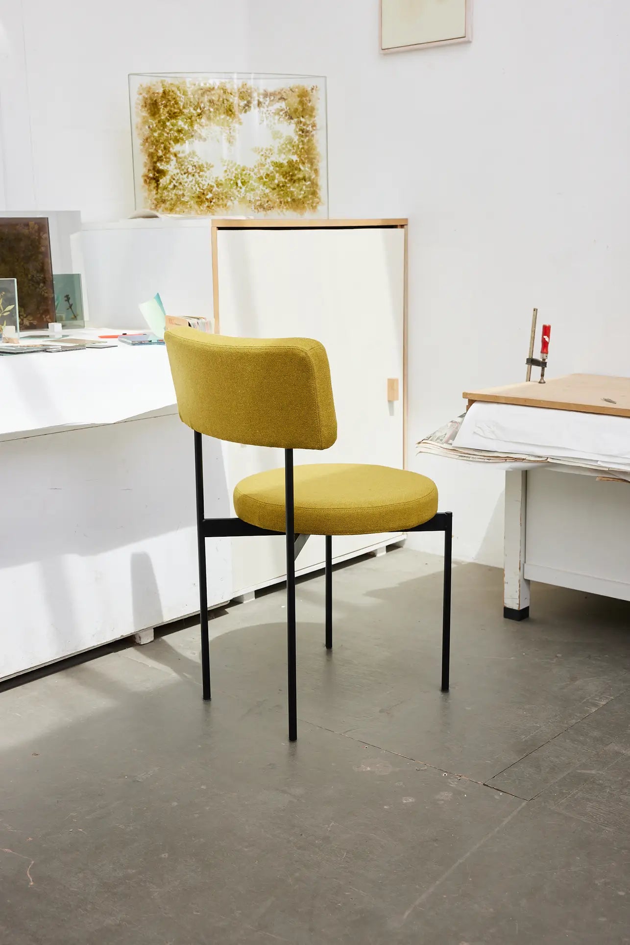 The HKLIVING Dining Chair, Main Line Flax in yellow with black metal legs by HKLIVING sits in a bright, minimalist room featuring white walls, scattered art supplies, and artwork across tables and surfaces.