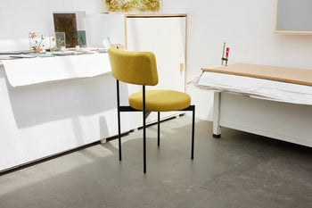 The HKLIVING Dining Chair, Main Line Flax, Black features a mustard-yellow cushioned seat and backrest with slim black metal legs, highlighted in a minimalist studio setting with white tables and background artwork, reflecting Dutch design by HKLIVING.