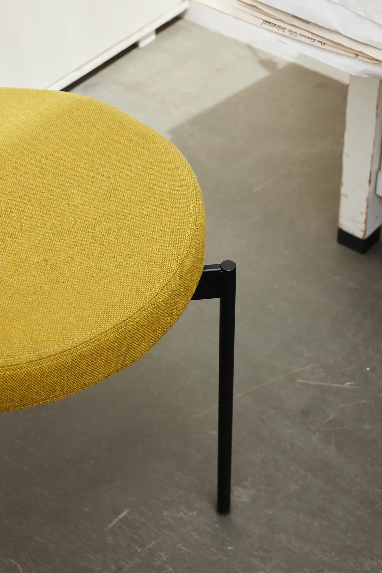 A close-up of the HKLIVING Dining Chair in Main Line Flax with a round, mustard-yellow seat and black metal legs, placed on a gray floor beside a white table.
