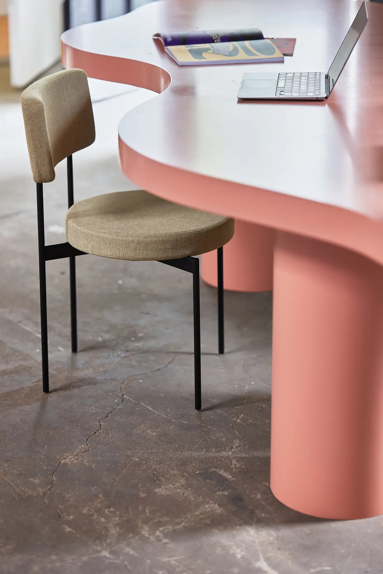 A modern workspace with a curved pink desk, an HKLIVING Dining Chair in Main Line Flax by HKLIVING featuring black metal legs, an open laptop, and a magazine on the tabletop. The floor is gray concrete with visible cracks.