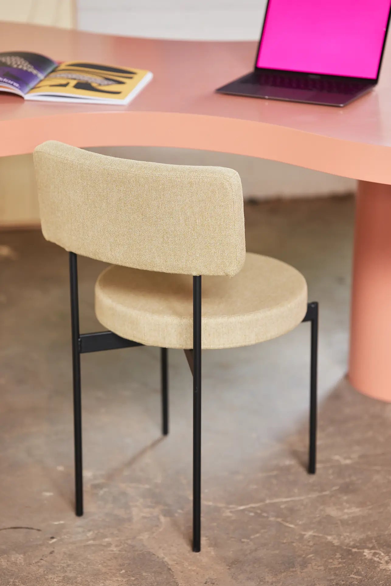 The HKLIVING Dining Chair, Main Line Flax in beige with black legs is set before a curved pink desk holding a laptop and open magazine on the concrete floor.