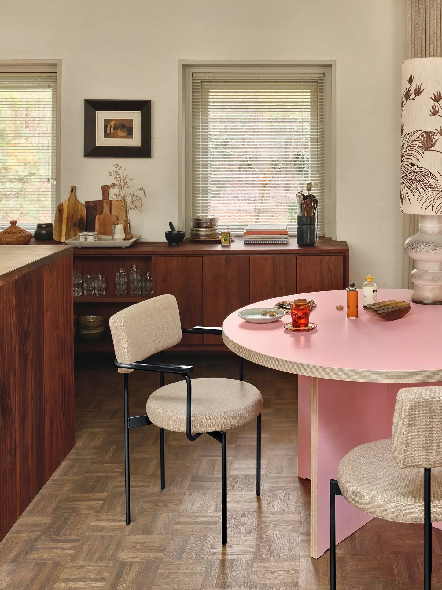 A cozy kitchen features wood cabinets, a round pink table with two HKLIVING Dining Chairs in Main Line Flax Black, decorative cups, vases, plants, windows with blinds for natural light, and framed art on the wall.