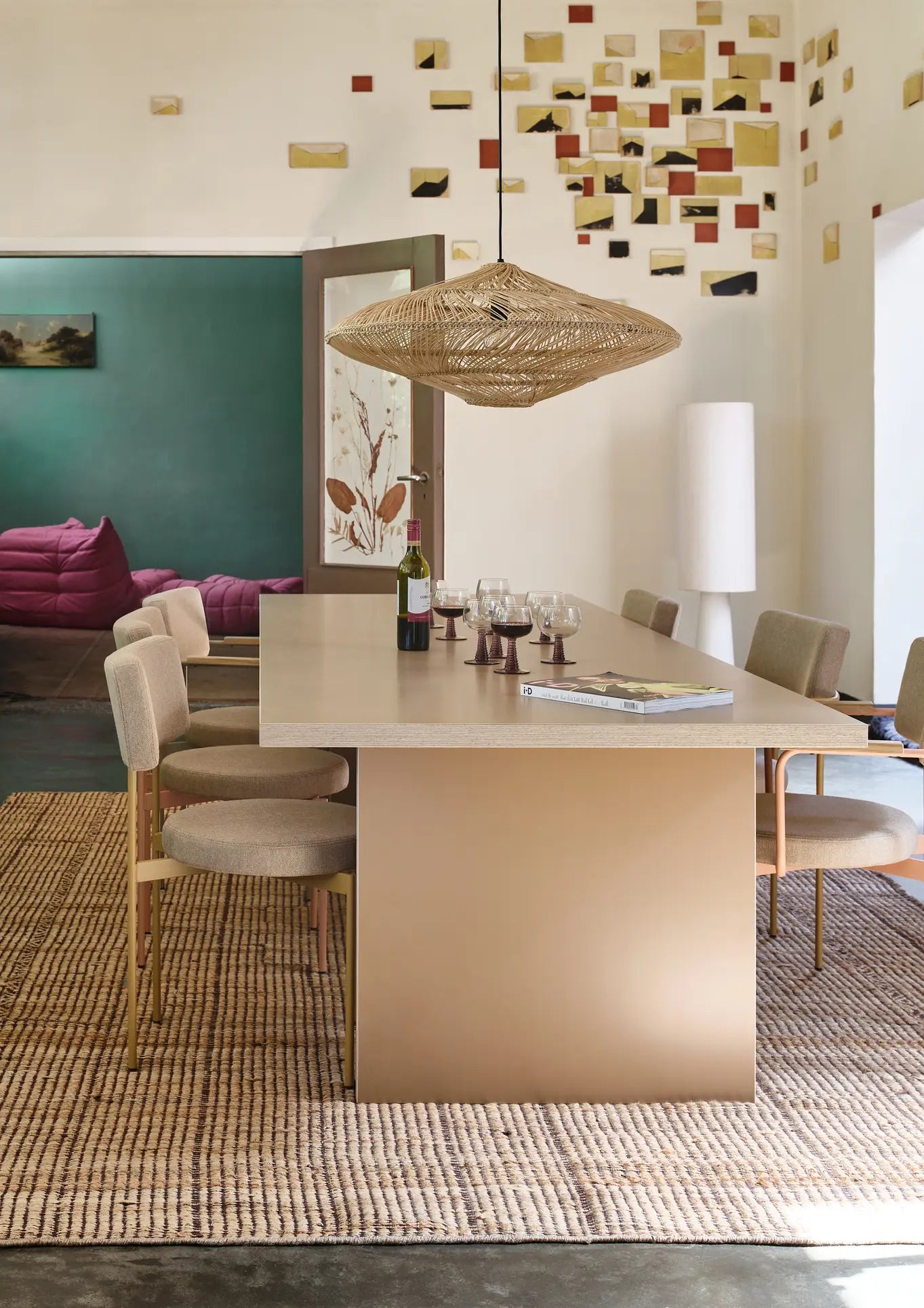 A modern dining room with a long beige table, six HKLIVING Dining Chairs in Main Line Flax Dusty, a woven pendant light, wine glasses and bottle on the table, plus a textured rug and colorful abstract wall art.