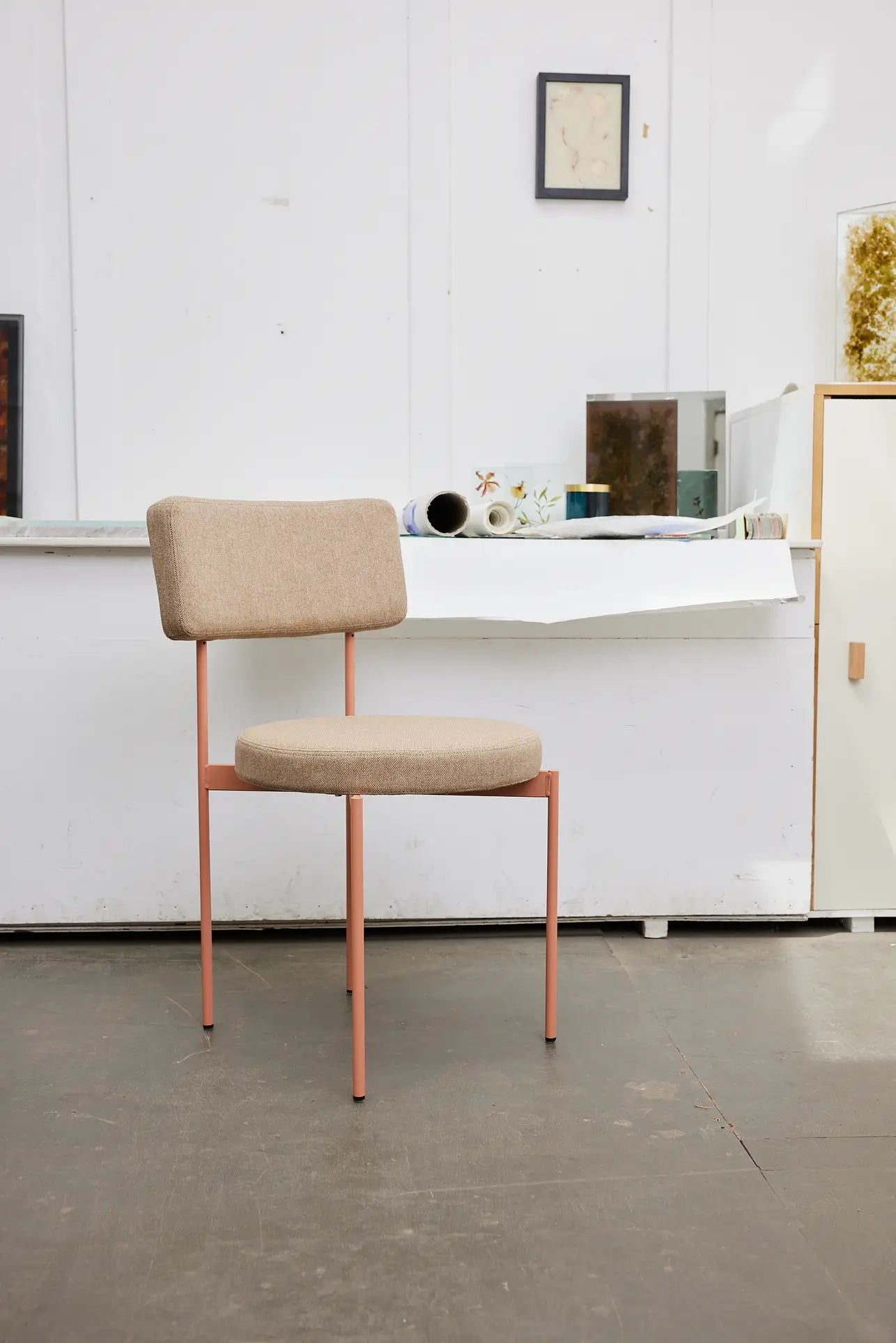 The HKLIVING Dining Chair, Main Line Flax, Nude from HKLIVING features a beige cushioned seat and slim pink metal legs, adding modern Dutch design to a minimalist room with white walls, gray floor, and a stylishly decorated counter.
