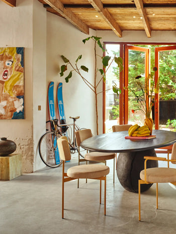 A modern dining room features a round black table, HKLIVING Dining Chair in Main Line Flax Nude, a bowl of bananas, and tall branches in a vase. Blue skis and a bicycle rest by the wall; sunlight fills the space with color and art.