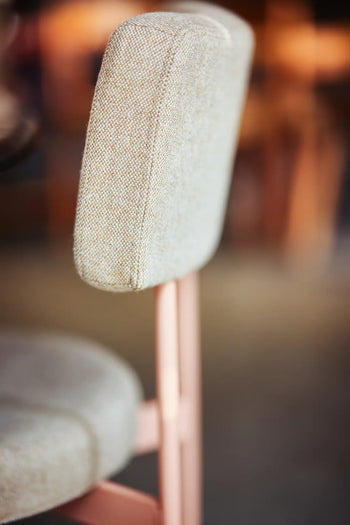 A close-up of the HKLIVING Dining Chair, Main Line Flax in Nude, featuring a light beige upholstered backrest and seat on a light pink metal frame, with a blurred background.