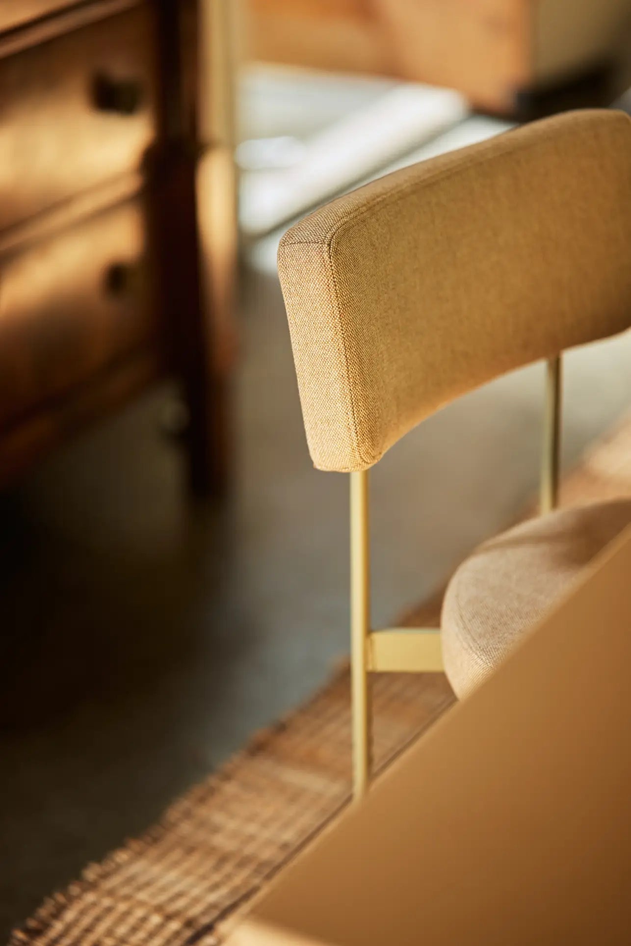 A close-up of the HKLIVING Dining Chair, Main Line Flax in Olive by HKLIVING, featuring beige upholstery and a metal frame, positioned near a woven rug and wooden cabinet in a softly lit room.