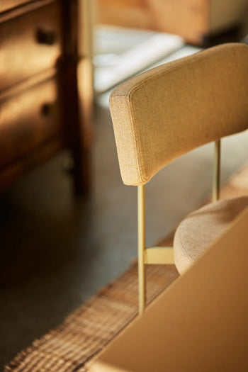 A close-up of the HKLIVING Dining Chair, Main Line Flax in Olive by HKLIVING, featuring beige upholstery and a metal frame, positioned near a woven rug and wooden cabinet in a softly lit room.