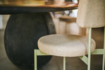 A close-up of the HKLIVING Dining Chair, Main Line Flax in Olive by HKLIVING, featuring a metal frame and round beige cushion near a dark, round-based table indoors. This customizable chair brings contemporary style to your space.