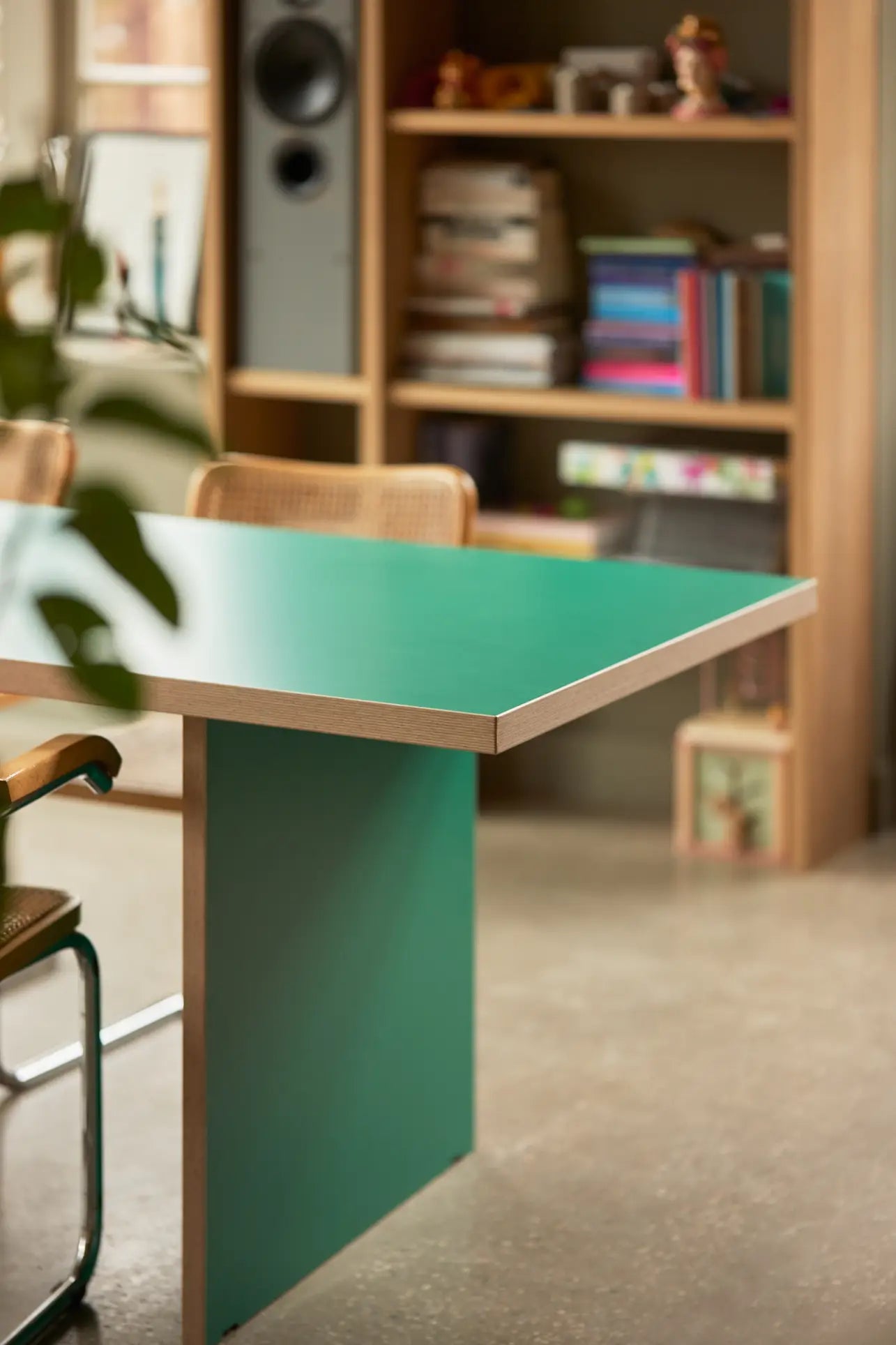 The HKLIVING Dining Table, Rectangular, 220cm from HKLIVING with a green surface and wooden edges is paired with chairs in a room featuring a book-filled bookshelf, concrete floor, and leafy plant for a modern stylish look.