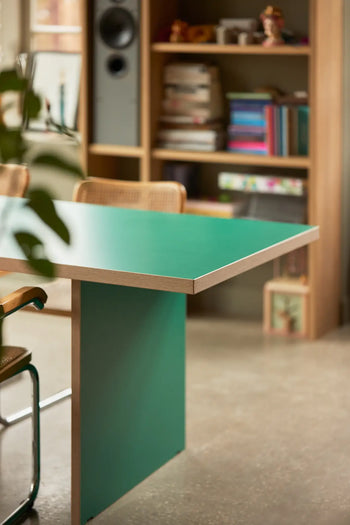 The HKLIVING Dining Table, Rectangular, 220cm from HKLIVING with a green surface and wooden edges is paired with chairs in a room featuring a book-filled bookshelf, concrete floor, and leafy plant for a modern stylish look.